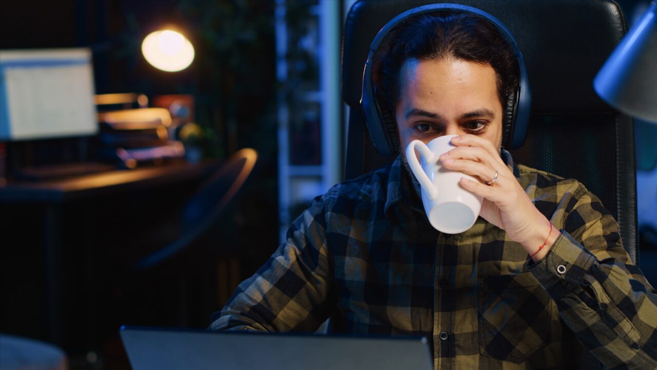 Mężczyzna w słuchawkach pije kawę przy biurku z laptopem.