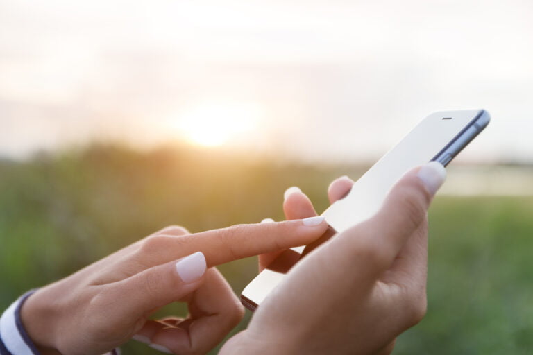 Dłoń trzymająca smartfon, druga dłoń dotykająca ekranu, rozmyte tło zewnętrzne w świetle zachodzącego słońca.