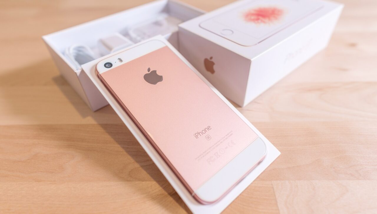 Różowy iPhone SE na opakowaniu, widoczne pudełko i akcesoria na stole.