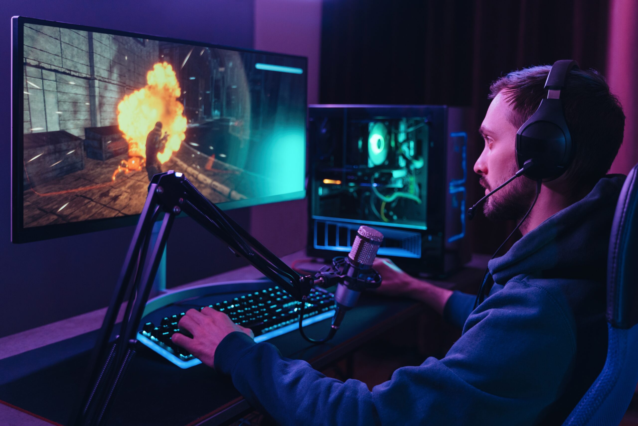 Osoba z zestawem słuchawkowym grająca w grę komputerową na monitorze, przy biurku z mikrofonem i nowoczesnym sprzętem komputerowym podświetlonym na niebiesko.