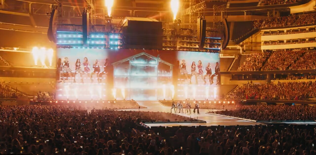 Koncert na stadionie z dużą ilością widzów, scena oświetlona, tancerze i wybuchy pirotechniczne na scenie.