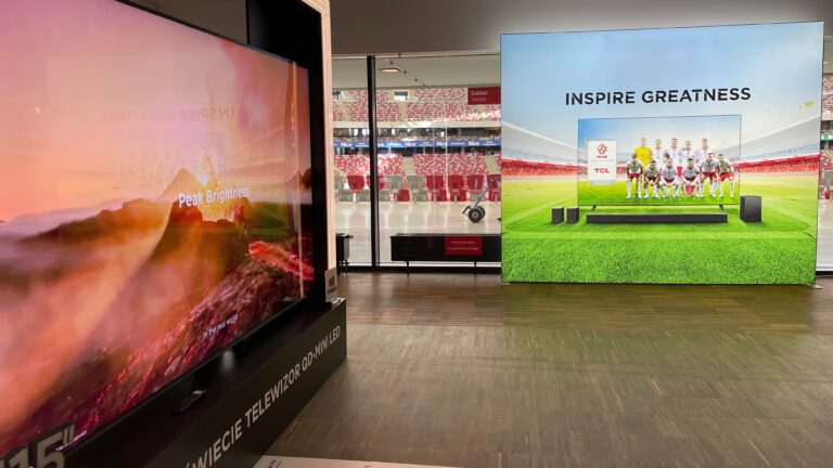 Wnętrze sklepu z telewizorami, z dużym, zaokrąglonym ekranem po lewej stronie i wielkim plakatem reklamowym z piłkarzami po prawej.