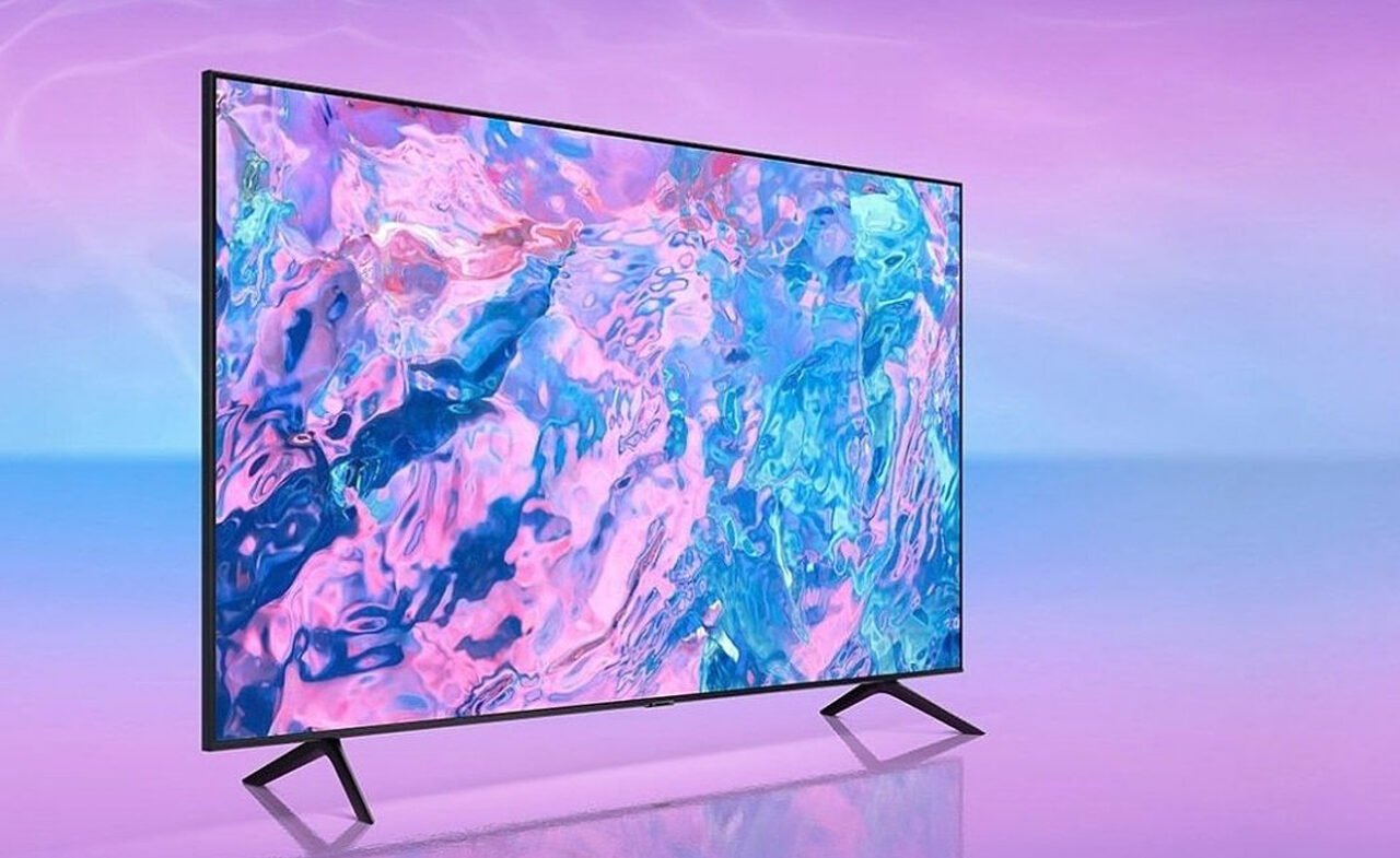 Duży telewizor z wyświetlaczem o żywych kolorach, stojący na dwóch nóżkach na tle abstrakcyjnego różowo-niebieskiego tła.