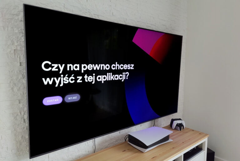 Duża plaska telewizja na ścianie wyświetlająca wiadomość w języku polskim "Czy na pewno chcesz wyjść z tej aplikacji?" z przyciskami wyboru, obok konsola do gier i kontroler na drewnianej szafce.