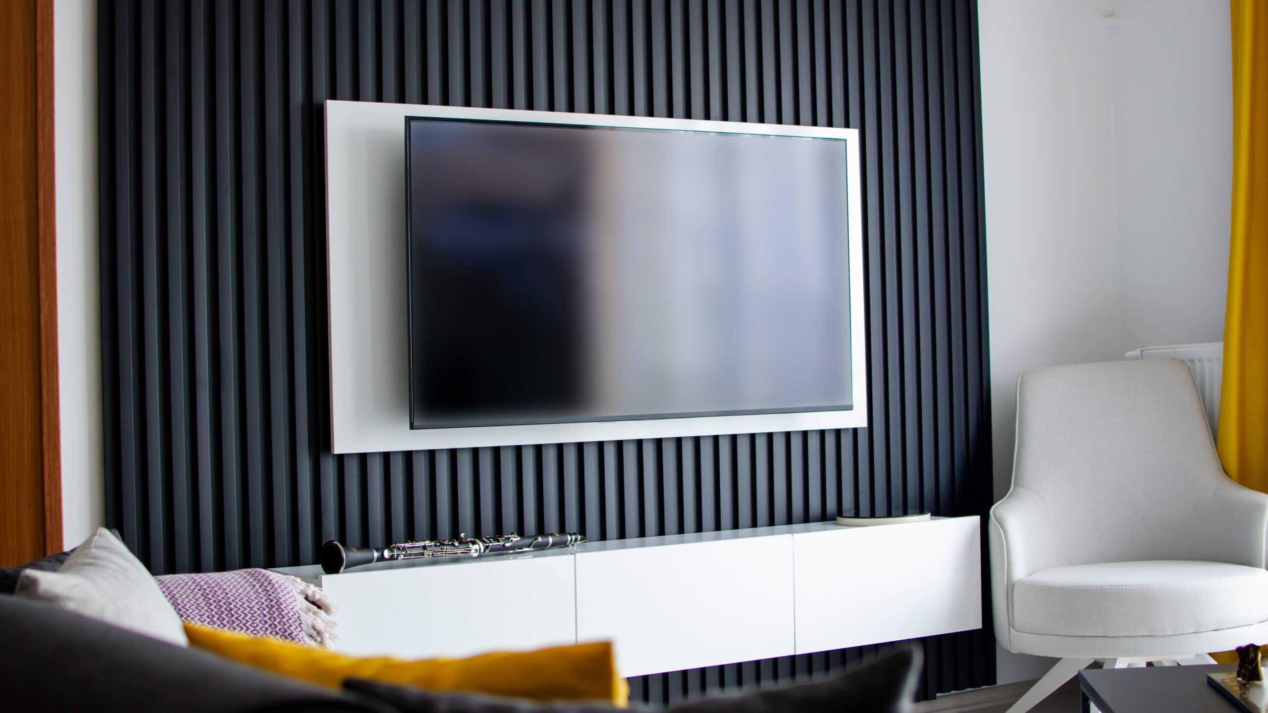 Współczesny salon z telewizorem na ścianie z czarnymi pionowymi panelami, białą szafką pod telewizorem, białym fotelem i żółtą zasłoną.