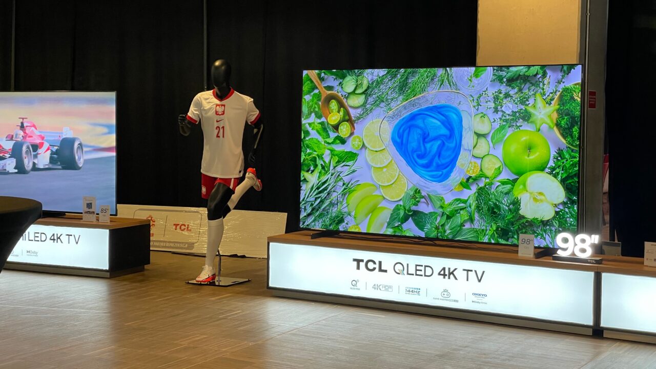 Dwa duże telewizory 4K z wyświetlanymi obrazami: po lewej wyścigówka Formuły 1, a po prawej owoce i zioła, między nimi manekin w białej koszulce i czerwonych spodenkach.