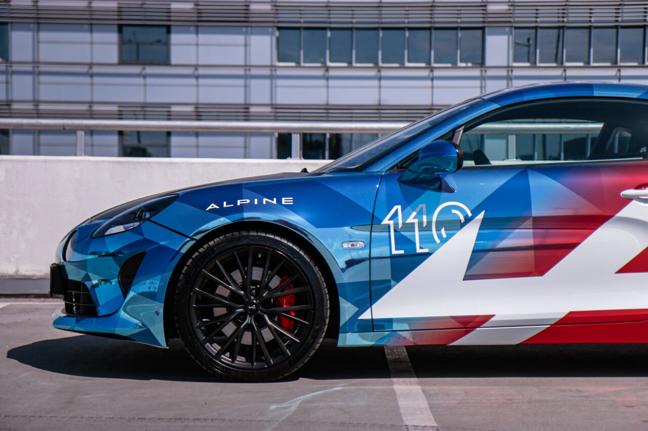 Test Alpine A110 S - bok niebieskiego sportowego samochodu z numerem 110 na tle nowoczesnego budynku.