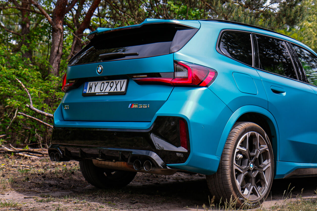 Tylny widok BMW X1 M35i zaparkowanego w lesie, widać charakterystyczne sportowe elementy i błękitny kolor karoserii.