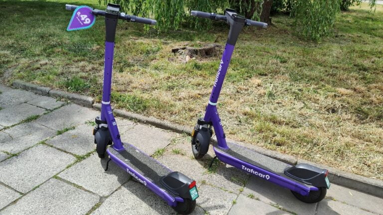 Dwa fioletowe hulajnogi elektryczne TrafiCar zaparkowane na chodniku obok trawnika.