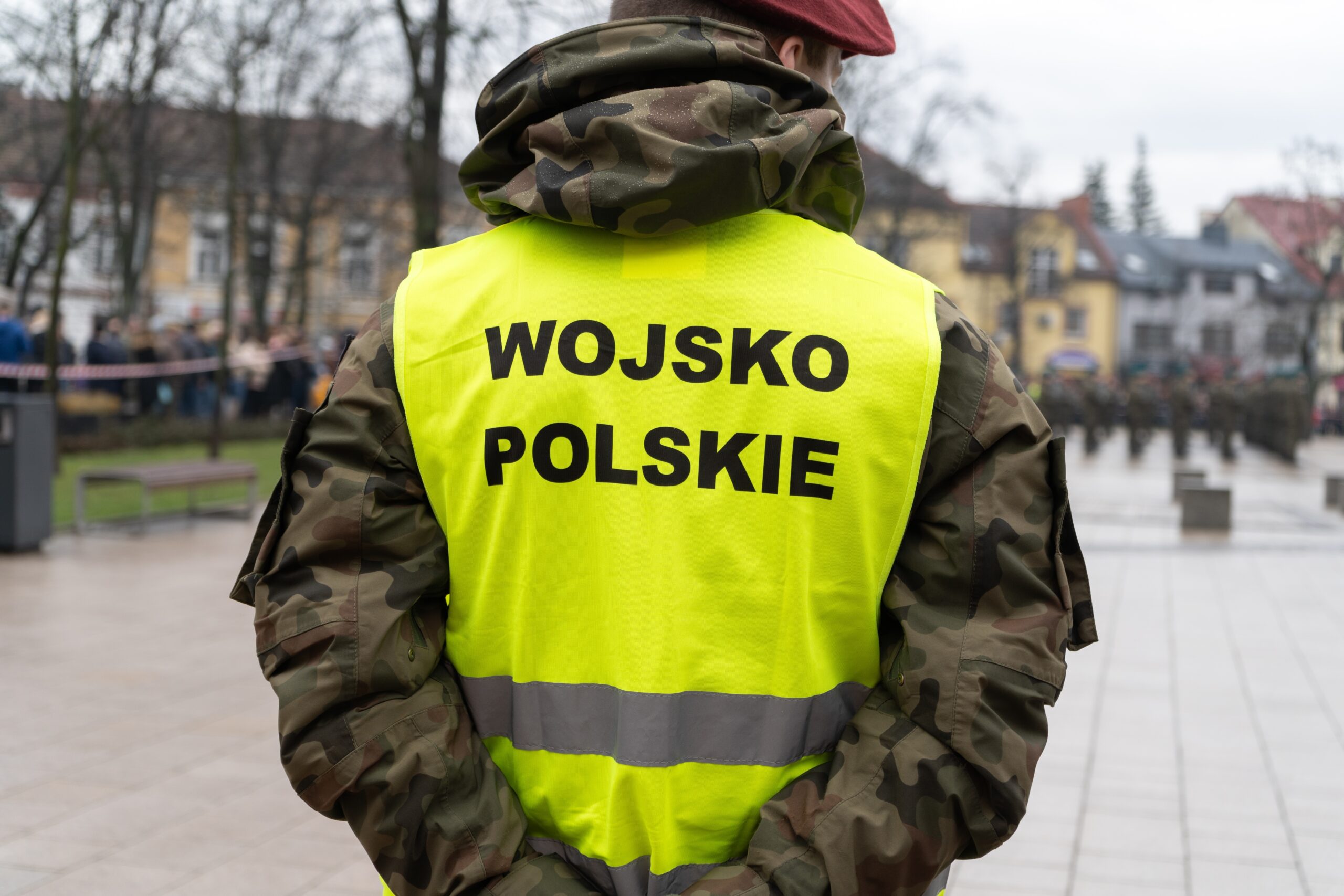 Żołnierz ubrany w kamuflaż i żółtą kamizelkę odblaskową z napisem "WOJSKO POLSKIE" stoi na placu miejskim.