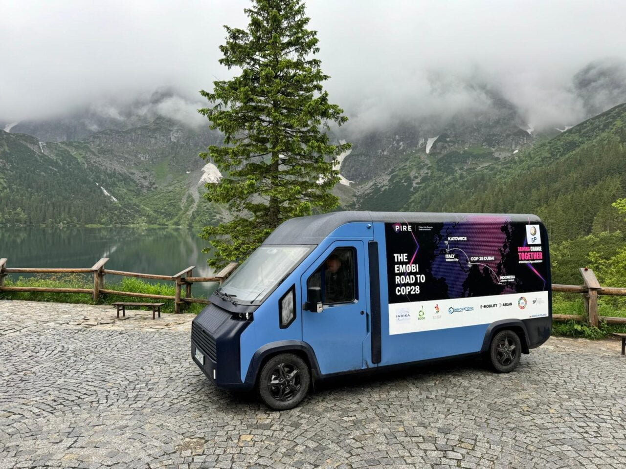 Elektryczny bus nad Morskie Oko Niebieska furgonetka z napisem "The Emobi Road to COP28" zaparkowana przy górskim jeziorze, w tle góry pokryte mgłą i lasem.