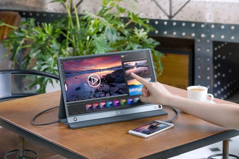 Osoba dotykająca ekranu dotykowego monitora ASUS z wyświetlonym edytorem zdjęć, na drewnianym stole obok filiżanki kawy i smartfona, w tle rośliny.