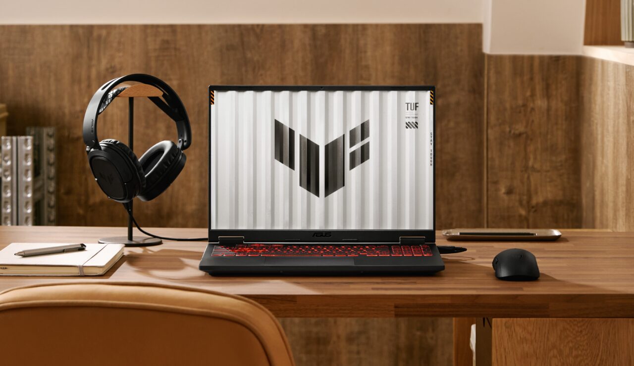 Laptop ASUS TUF na drewnianym biurku, z lewej strony stojak na słuchawki, notes i długopis, z prawej strony myszka i telefon komórkowy.