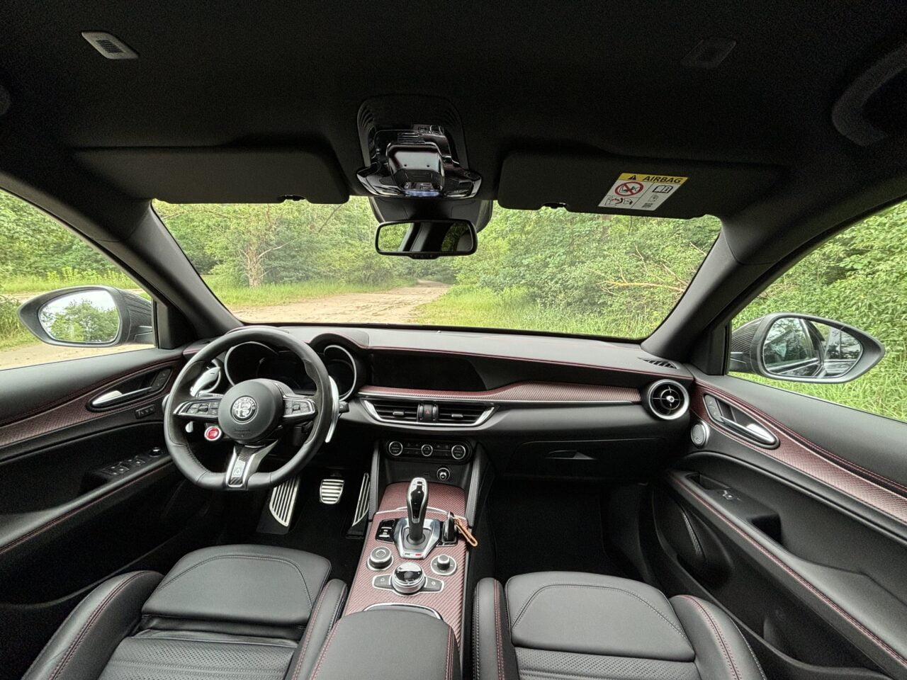 Wnętrze samochodu marki Alfa Romeo z widokiem na kierownicę, konsolę środkową i deskę rozdzielczą, na tle widoczna ścieżka w lesie.