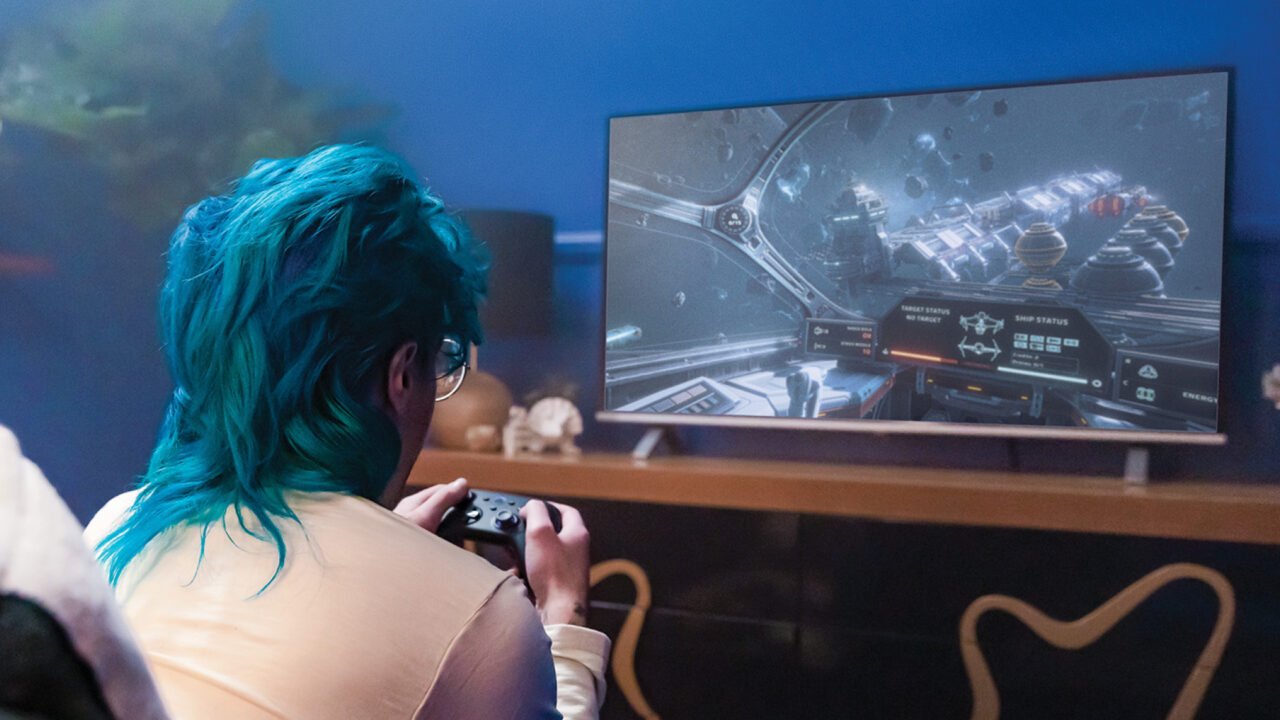 Osoba z niebieskimi włosami grająca w grę komputerową na dużym ekranie, trzymając kontroler.