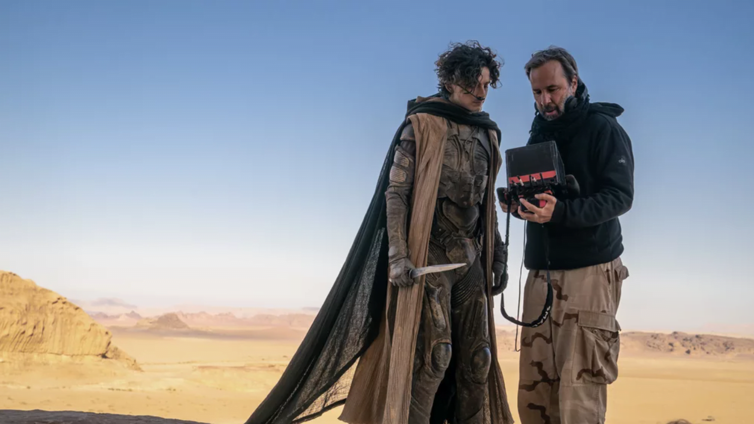 Aktor w kostiumie stoi na pustyni obok reżysera oglądającego ekran.