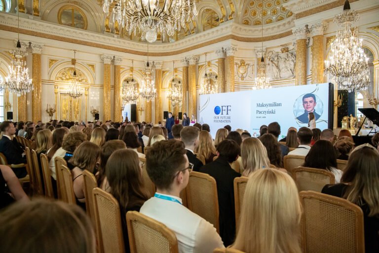 Zgromadzenie ludzi uczestniczących w konferencji w eleganckiej, złotej sali z żyrandolami, na scenie znajduje się ekran z napisem "Our Future Foundation" i zdjęciem młodego mężczyzny.