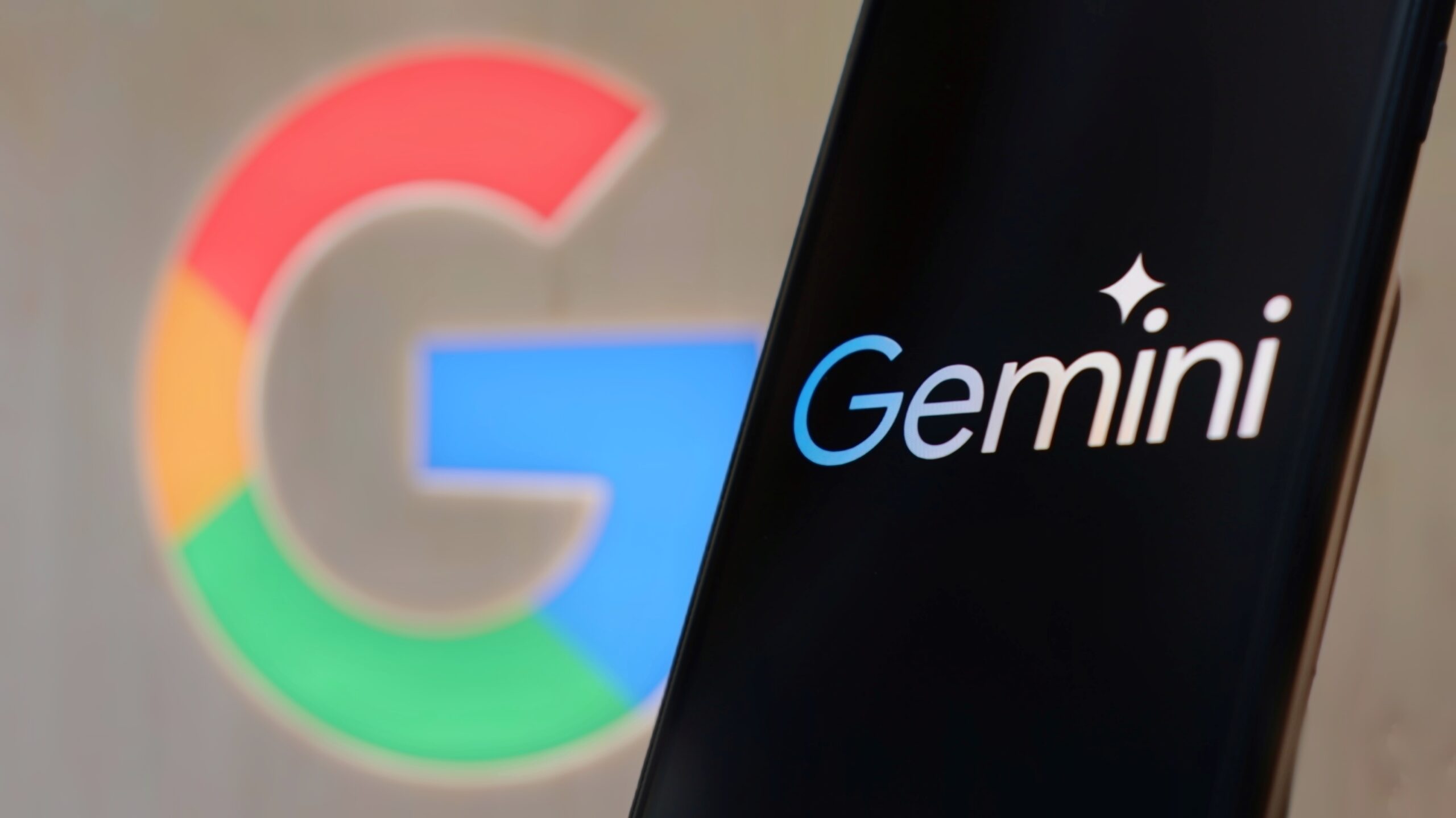 Logo Google w tle, napis "Gemini" na ekranie smartfona na pierwszym planie.