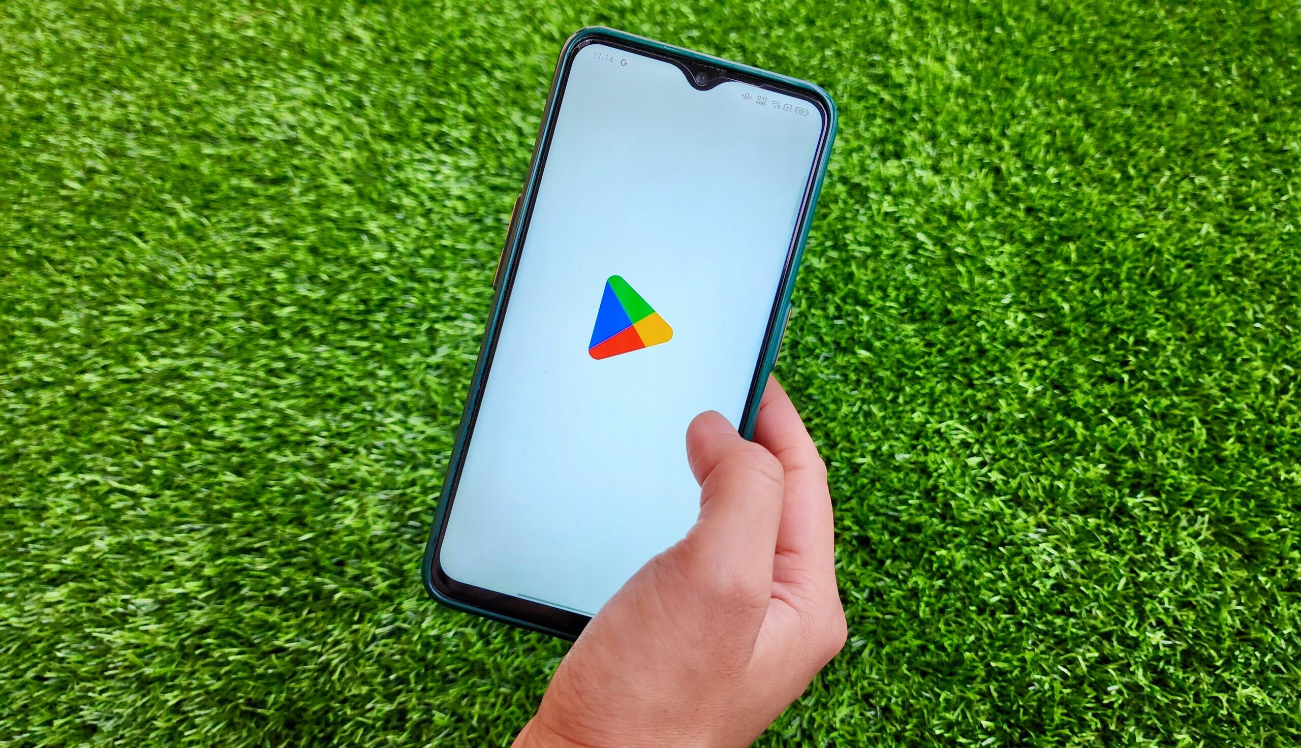 Dłoń trzymająca smartfon z ekranem pokazującym logo Google Play na tle zielonej trawy.
