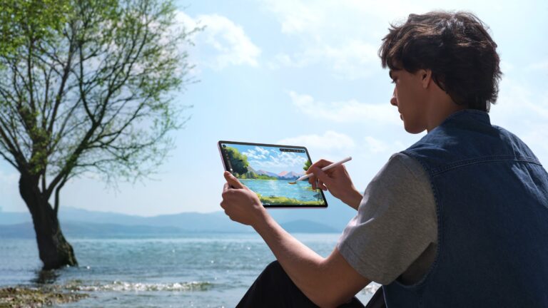 Osoba trzymająca tablet, rysująca pejzaż za pomocą pióra cyfrowego nad jeziorem, z drzewem i górami w tle.