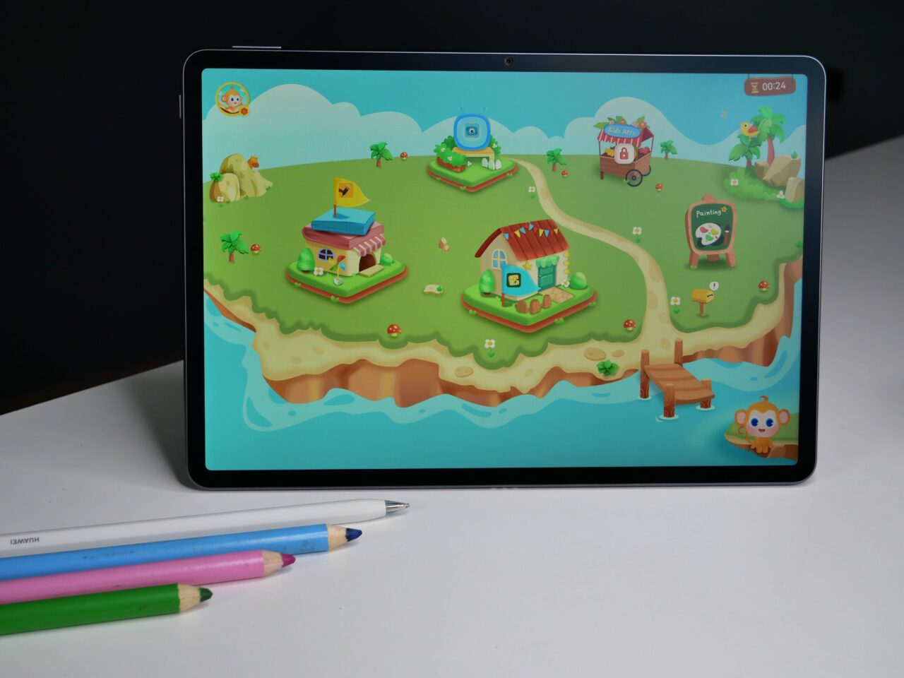 Tablet na białym stole z wyświetlonym kolorowym interfejsem gry edukacyjnej dla dzieci oraz leżące obok kolorowe kredki.