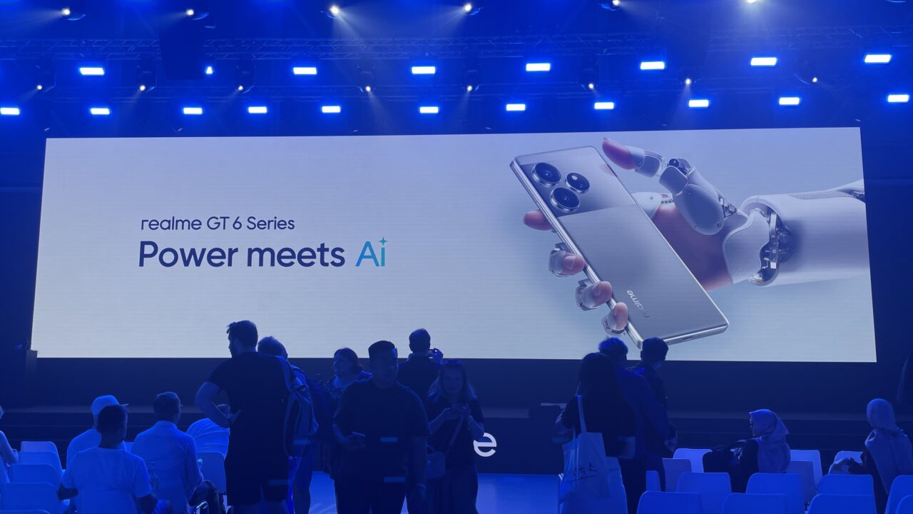 Osoby na tle ekranu z napisem "realme GT 6 Series Power meets Ai" i obrazkiem robota trzymającego smartfon.