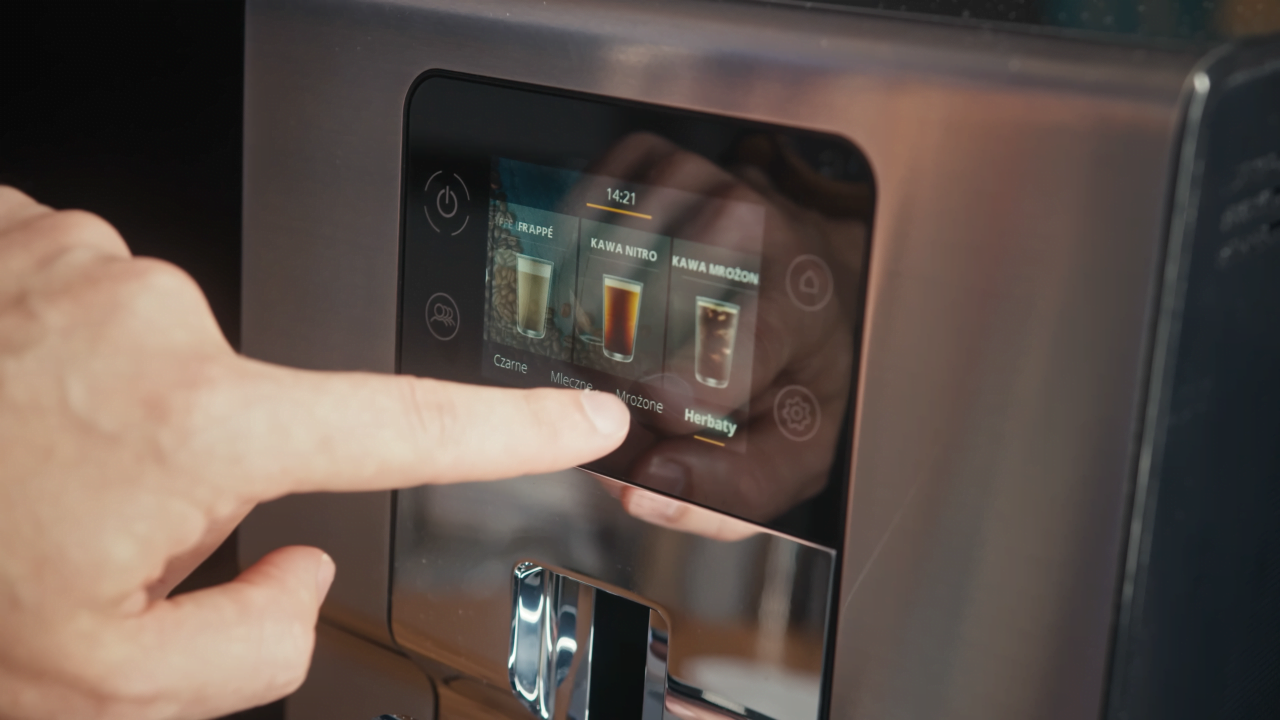 Palec dotykający ekran dotykowy ekspresu do kawy, wyświetlający różne opcje napojów.