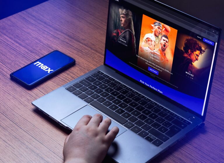 Osoba trzymająca rękę na touchpadzie laptopa, na ekranie otwarta strona z ofertą serwisu streamingowego Max; obok leży telefon z logo Max na wyświetlaczu.