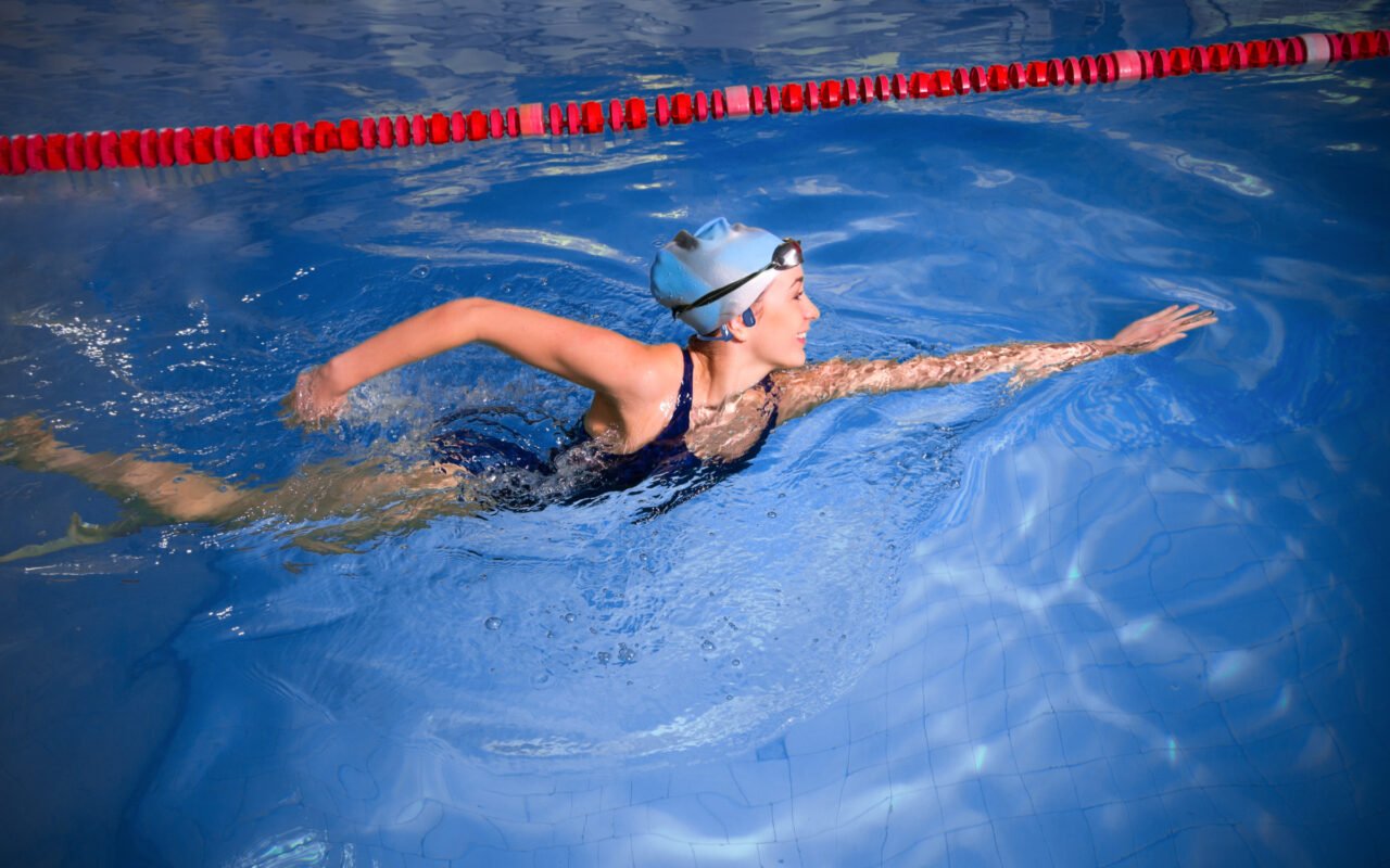 Oto słuchawki dla sportowców od OPN SOUND. Dziewczyna w czepku pływackim płynąca w basenie stylem grzbietowym obok boi oddzielającej tory.