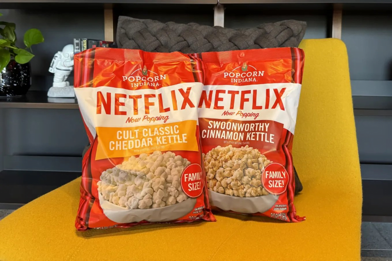 Dwie torby popcornu marki Popcorn Indiana z napisem Netflix na żółtym krześle; smaki Cult Classic Cheddar Kettle i Swoonworthy Cinnamon Kettle.