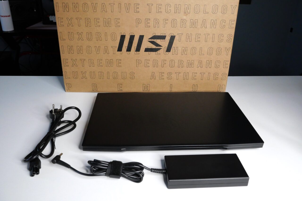Laptop MSI z kablami zasilania i ładowarką na białym stole, w tle karton z napisami.