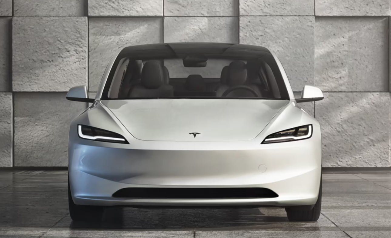Biały samochód Tesla Model 3 z widokiem od frontu na tle kamiennej ściany.