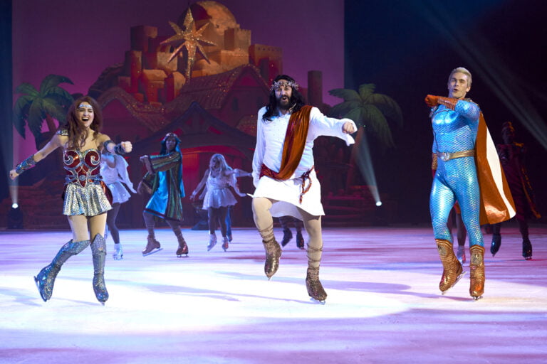 Kadr z The Boys 4. Artyści w kolorowych kostiumach (w tym jedna osoba przebrana za superbohaterkę, druga za Jezusa, a trzecia za superbohatera w niebieskim stroju) występują na lodzie w scenerii z palmami i budynkami w tle.