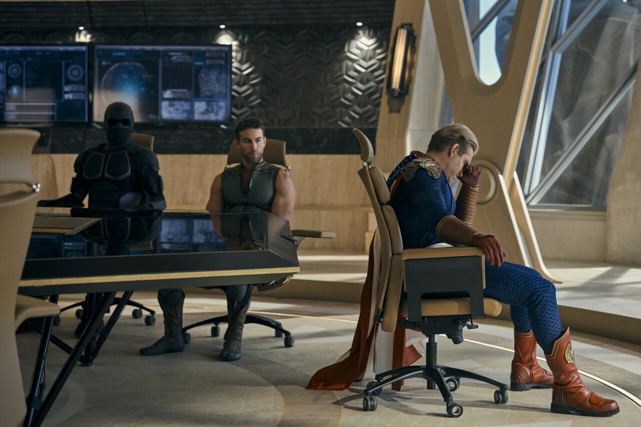 Trzech mężczyzn siedzących przy nowoczesnym stole w jasno oświetlonym pokoju, jeden z nich trzyma się za głowę, wyglądając na zmartwionego.