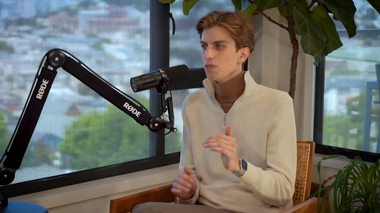 Mężczyzna rozmawiający do mikrofonu RODE podczas nagrywania podcastu, siedząc na krześle obok okna i rośliny, w jasnym swetrze z golfem.