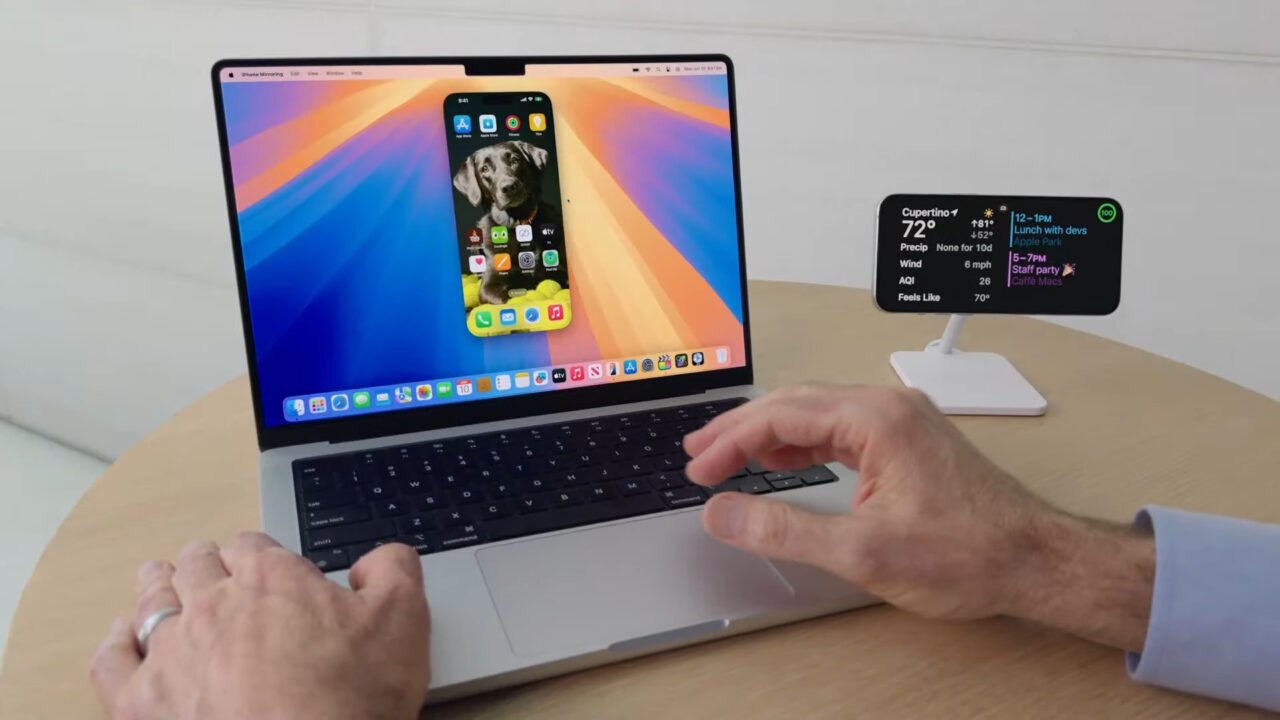 Osoba obsługująca laptopa z funkcją lustrzanego odbicia ekranu iPhone'a; obok stoi stojak z ekranem pokazującym pogodę i kalendarz.