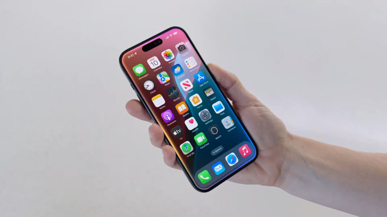 iOS 18. Telefon komórkowy trzymany w dłoni, wyświetlający ekran główny z ikonami aplikacji.