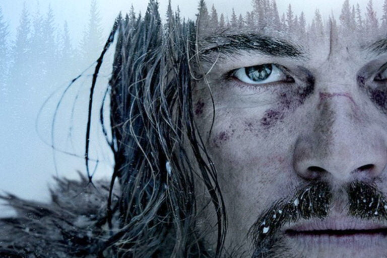 Platakt do filmu Zjawa. Zbliżenie twarzy mężczyzny w zimowej scenerii, z brodą i posiniaczoną twarzą, w tle widoczne zarysy drzew ospadanych śniegiem.