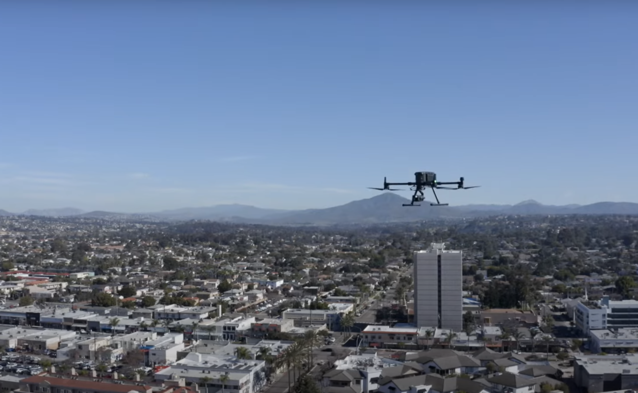Dron unoszący się nad miastem z widokiem na budynki, ulice i góry w tle.