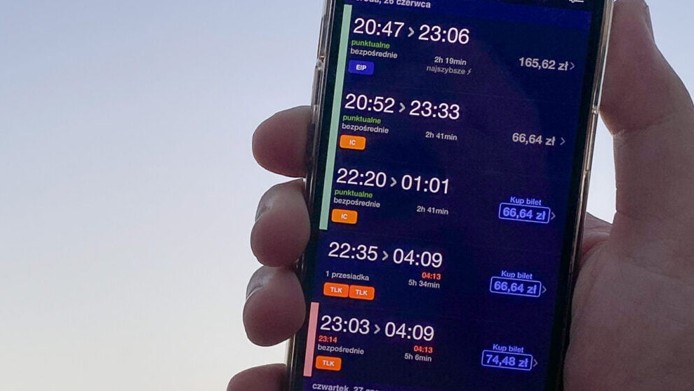 Opóźnienie pociągu w aplikacji KOLEO - Telefon trzymany w dłoni, na ekranie wyświetlane są informacje o rozkładzie jazdy pociągów w języku polskim.