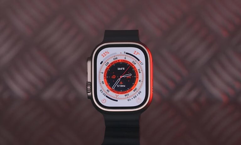 Zbliżenie na inteligentny zegarek z pomarańczowym pierścieniem i czarnym paskiem, wyświetlający tarczę z danymi nawigacyjnymi i godziną 5:13 PM.