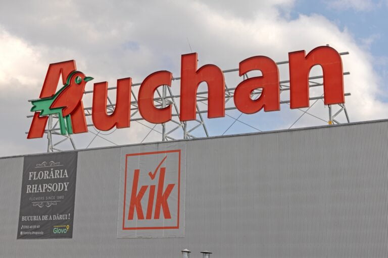 Znak Auchan z czerwonym napisem i zielonym symbolem ptaka oraz znak KiK na budynku. Baner reklamy kwiaciarni Florăria Rhapsody po lewej stronie.