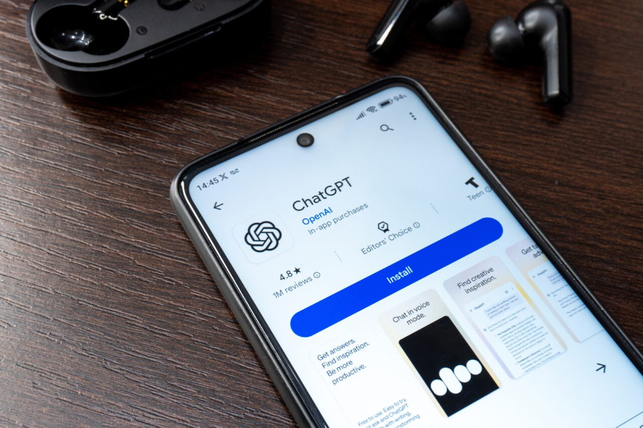 Ekran smartfona z wyświetlonym ekranem pobierania aplikacji ChatGPT na Google Play, obok czarne bezprzewodowe słuchawki na drewnianym stole.