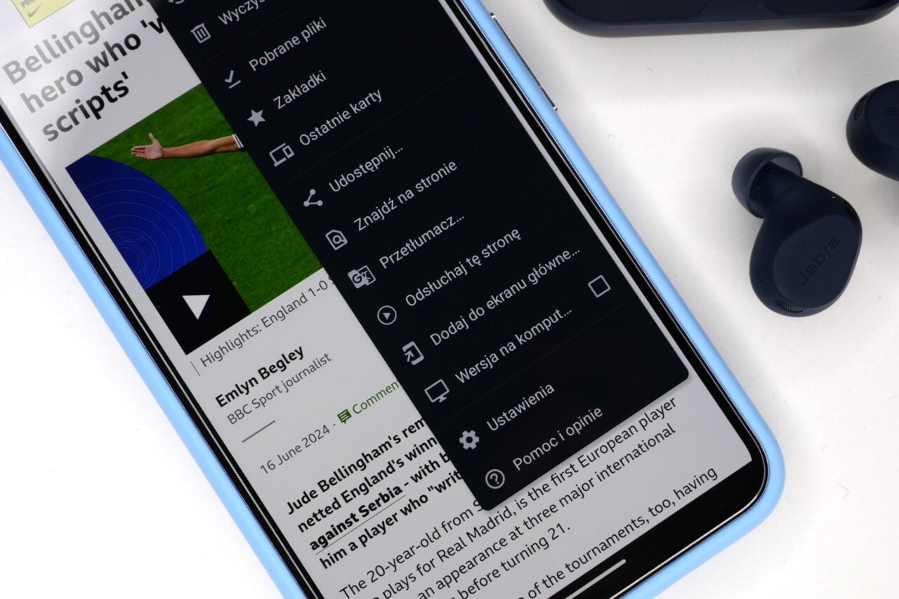 Smartfon z otwartą stroną internetową oraz menu przeglądarki, obok leżą słuchawki douszne Jabra.