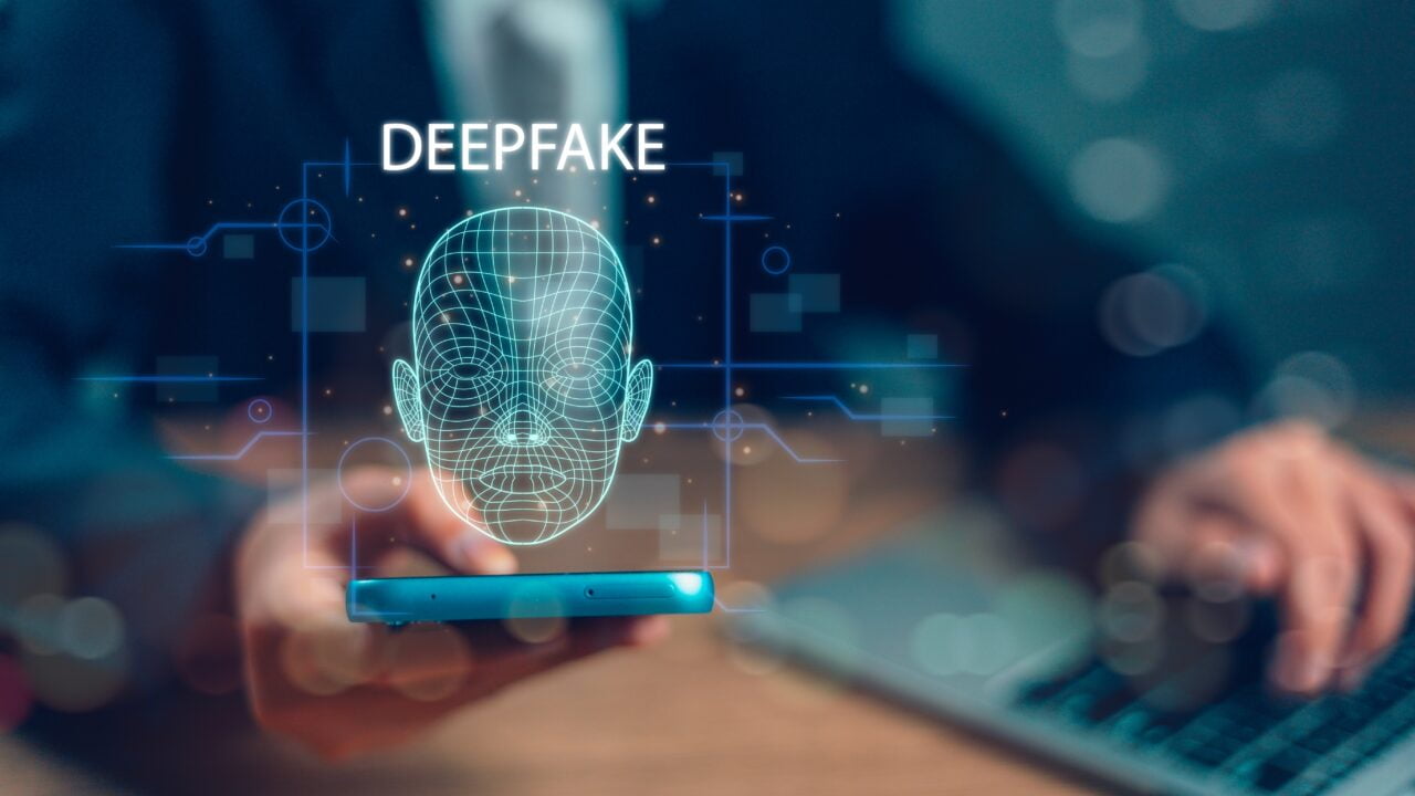 Osoba trzymająca telefon z wyświetlaną grafiką siatki 3D twarzy i napisem deepfake