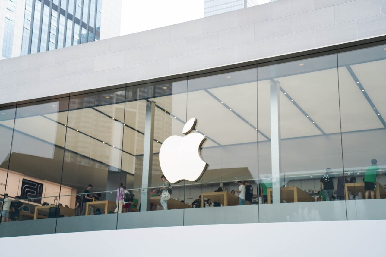 Apple Store z dużym logotypem jabłka widocznym na szklanej fasadzie. Wewnątrz sklepu widać klientów i pracowników.