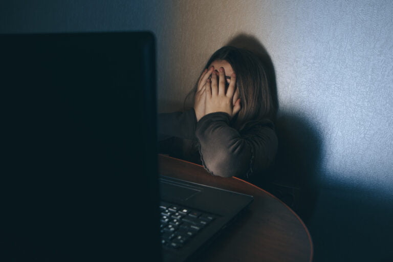 Osoba siedząca przed laptopem, zakrywająca twarz rękami.