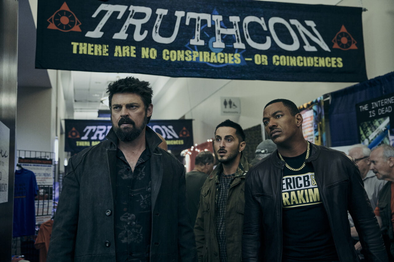 Gdzie obejrzeć The Boys - kadr z serialu. Trzej mężczyźni stojący przed banerem z napisem "TRUTHCON" na wydarzeniu o teoriach spiskowych.
