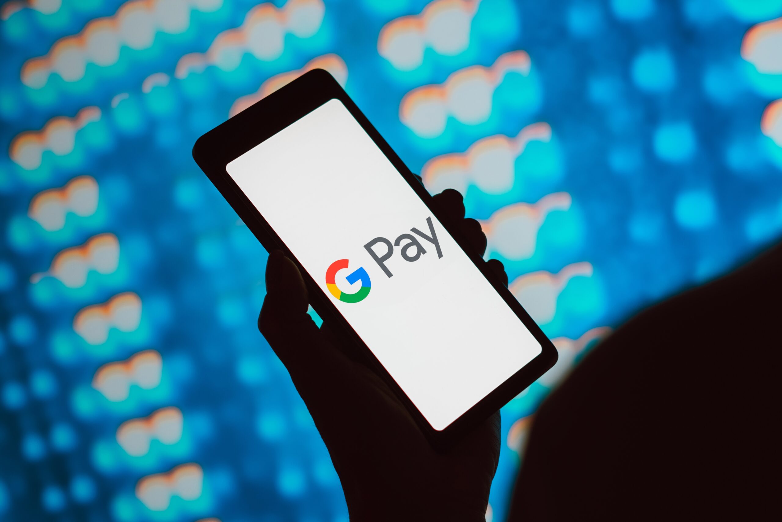 Osoba trzymająca telefon z logo Google Pay na ekranie, w tle rozmazane, kolorowe wzory.