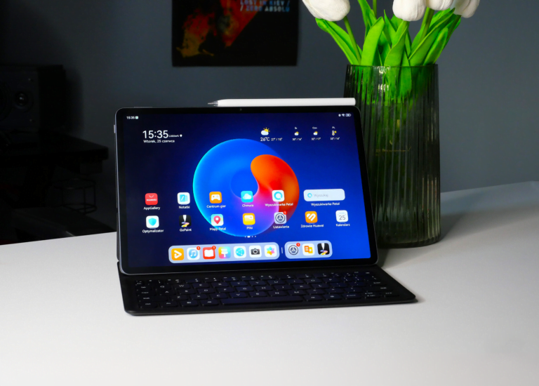 Tablet z klawiaturą na biurku, obok wazonu z kwiatami. Na ekranie wyświetlony ekran główny z ikonami aplikacji.